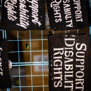 May include: Black fabric squares with white text that reads "Support Disability Rights." The squares are hanging on a blue grid. The text is in a handwritten style. The image is well-lit and the text is easy to read.