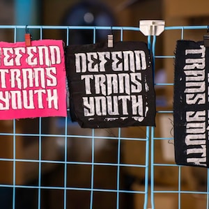 May include: Three fabric patches with the text "DEFEND TRANS YOUTH" in white block letters. One patch is hot pink, one is black, and the third is black with the text running vertically. The patches are clipped to a blue wire grid.