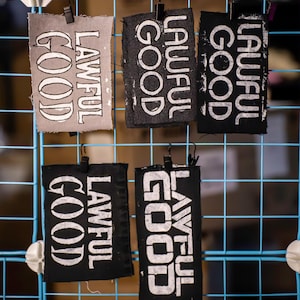 May include: A display of fabric tags with the words "Lawful Good" printed in white. The tags are in various shades of gray and black, hanging on a blue wire grid. The text is in a bold, sans-serif font.