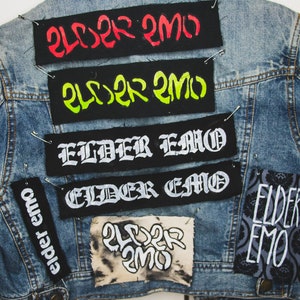 May include: A denim jacket with multiple patches sewn on. The patches are black with white text that says "Elder Emo" in different fonts and styles. Some patches have the text in red and green.