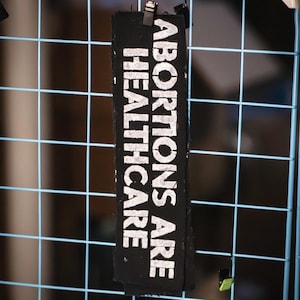 May include: A black rectangular fabric with the words "ABORTIONS ARE HEALTHCARE" printed in white. The fabric is hanging on a blue wire grid. The text is vertically oriented and centered on the fabric.