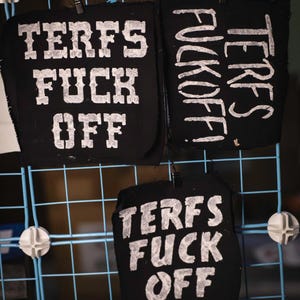 May include: Black fabric patches with white text that reads "TERFS FUCK OFF" are displayed on a blue grid. The patches are attached with small black clips. The text is in a bold, blocky font.