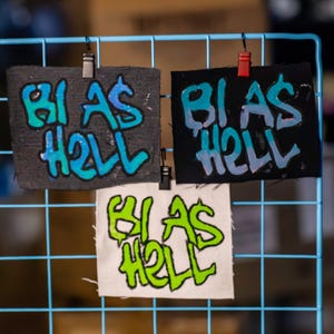 May include: Three fabric patches with the phrase "BI AS HELL" in graffiti-style lettering. The patches are attached to a blue grid. The top left patch is dark gray with blue and green text. The top right patch is black with blue and light blue text. The bottom patch is white with green text.