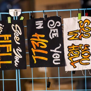 May include: Three handmade signs with the phrases "See You in Hell" and "In Hell" written in white and orange lettering on black fabric and white fabric. The signs are clipped to a blue wire grid.