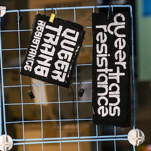May include: Two black fabric patches with white text are displayed on a light blue wire grid. One patch reads "QUEER TRANS RESISTANCE" and the other reads "queer trans resistance". The patches are held in place with small black clips and a yellow clip.
