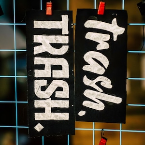 May include: Two black rectangular signs with the word "TRASH" in large, white, textured lettering. The signs are attached to a blue grid with red clips. The word "Trash" is written in a bold, artistic font.