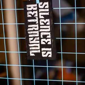 May include: A black rectangular patch with the words "SILENCE IS BETRAYAL" printed in white, hanging from a blue wire grid. The patch is attached with a small blue clip.