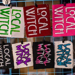 May include: A collection of fabric patches with the words "LOCAL WITCH" in various colors and styles. The patches are attached to a blue wire grid with small black clips. Colors include white, green, red, pink, black, and purple.