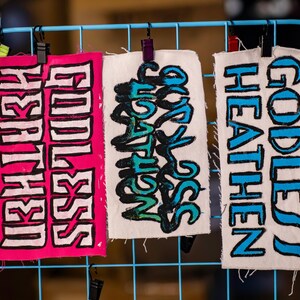 May include: Three fabric banners with bold text hang from a blue wire grid. The banners display the words "Goddess Earth", "Our Salvation", and "Goddess Heathen" in vibrant colors. The banners are held by black clips.