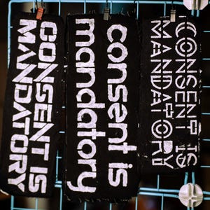 May include: Three black fabric patches with the words "CONSENT IS MANDATORY" in white block letters. The patches are hanging from a blue grid, held by small clips. The text is stenciled on the fabric.