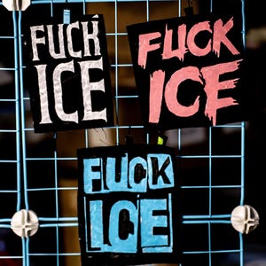 May include: Three black square signs with the words "FUCK ICE" in bold, stylized lettering. The text is white, pink, and blue. The signs are hanging on a blue wire grid.