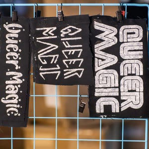 May include: Three black fabric patches with the words "Queer Magic" in white lettering. The patches are hanging on a blue wire grid. The text is in a bold, stylized font, with one patch featuring stars.
