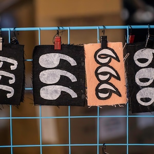 May include: Four fabric tags with the number "666" printed in white. The tags are black and peach, and are clipped to a blue wire grid. The tags are hanging from black clips and a red clip.
