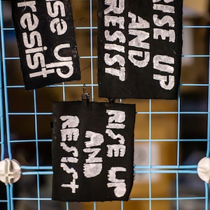 May include: Three black fabric signs with white text hanging on a blue wire grid. The signs read "Rise Up & Resist" and "Rise Up and Resist". The signs are held in place with small metal clips.