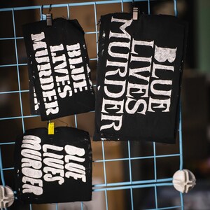 May include: Three black fabric squares with the words "BLUE LIVES MURDER" printed in white. The squares are clipped to a blue wire grid. The text is in a bold, sans-serif font.