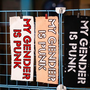 May include: Rectangular fabric patches with the text "MY GENDER IS PUNK" in red and white lettering. The patches are displayed on a blue wire grid, held by black clips. The fabric colors vary.