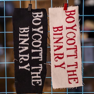 May include: Two rectangular fabric patches with the text "BOYCOTT THE BINARY". One patch is black with white text, and the other is off-white with red text. Both are hanging on a blue grid.