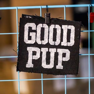 May include: A black fabric patch with the words "GOOD PUP" in large, white, block letters. The patch is attached to a blue wire grid with a black clip. The background is blurred.