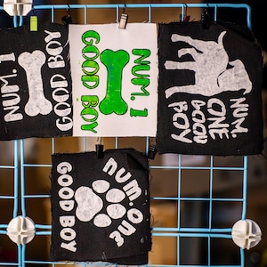 May include: A display of dog-themed patches. The patches are black and white, with the words "NUM. 1 GOOD BOY" and a bone graphic. Another patch has a dog silhouette, and a third has a paw print. They are clipped to a blue wire grid.