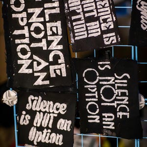 May include: Black fabric patches with white text that reads "Silence is NOT an Option" and "Silence is Not an Option". The patches are displayed on a blue grid.