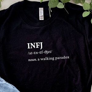 May include: Black t-shirt with white text that reads "INFJ /al-en-ef-dzer/ noun. a walking paradox". The t-shirt is laying on a white surface with a pair of blue jeans and a straw hat.