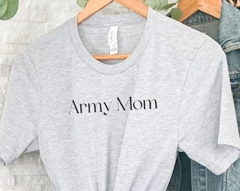 Könnte beinhalten: Ein graues T-Shirt mit dem Aufdruck "Army Mom" auf der Vorderseite. Das Shirt hängt an einem Kleiderständer mit einer Jeansjacke und einem gemusterten Rucksack.