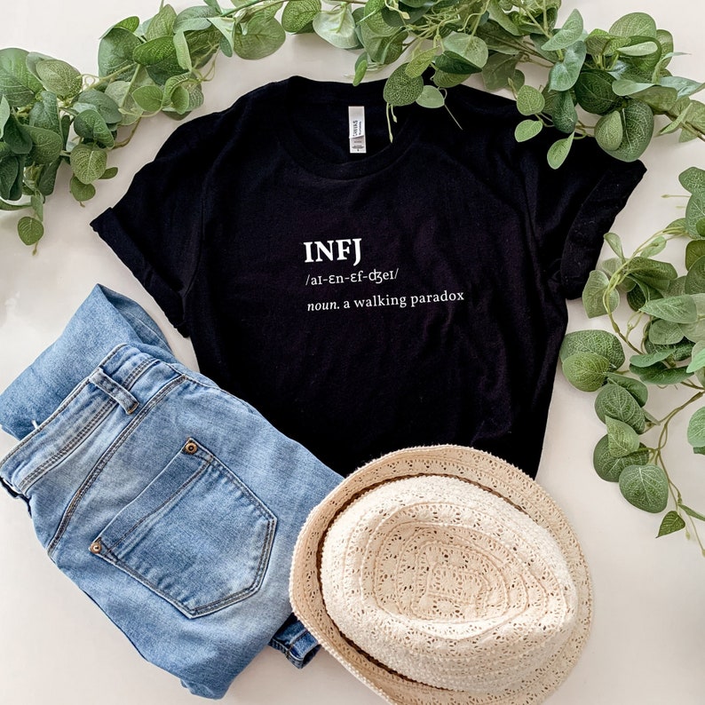 May include: Black t-shirt with white text that reads "INFJ /al-en-ef-dzer/ noun. a walking paradox". The t-shirt is laying on a white surface with a pair of blue jeans and a straw hat.