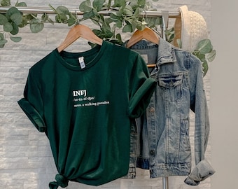 May include: A dark green t-shirt with white text that reads "INFJ /ˈɪn.f.dʒi/ noun. a walking paradox". The t-shirt is hanging on a rack with a denim jacket.
