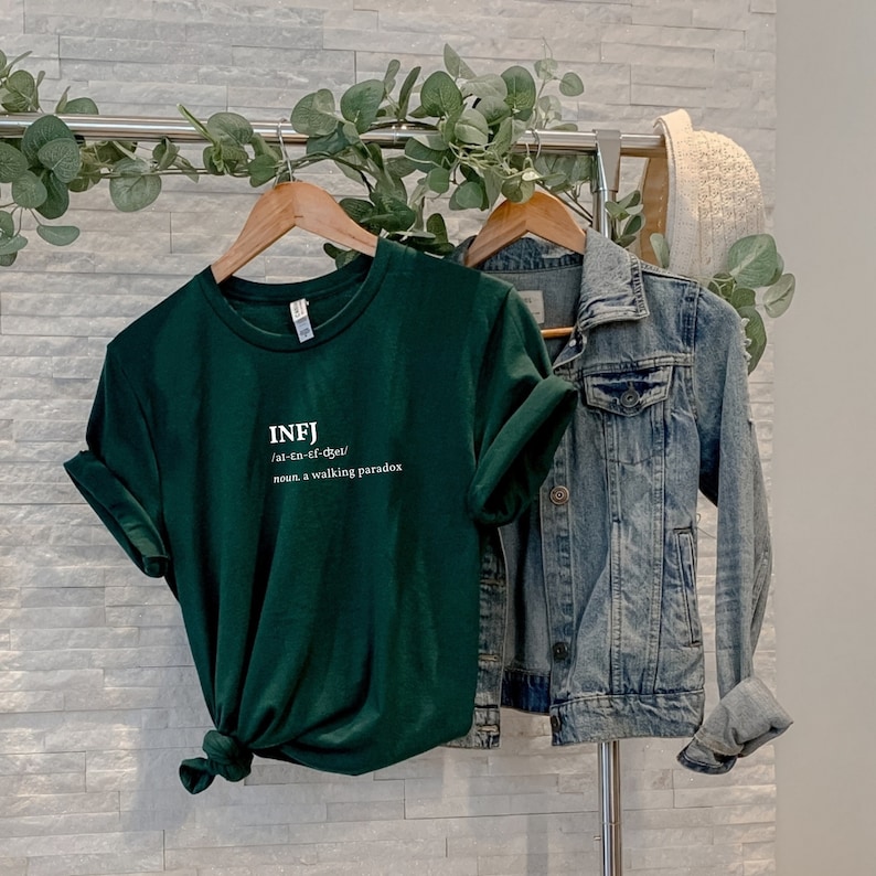 May include: A dark green t-shirt with white text that reads "INFJ /ˈɪn.f.dʒi/ noun. a walking paradox". The t-shirt is hanging on a rack with a denim jacket.