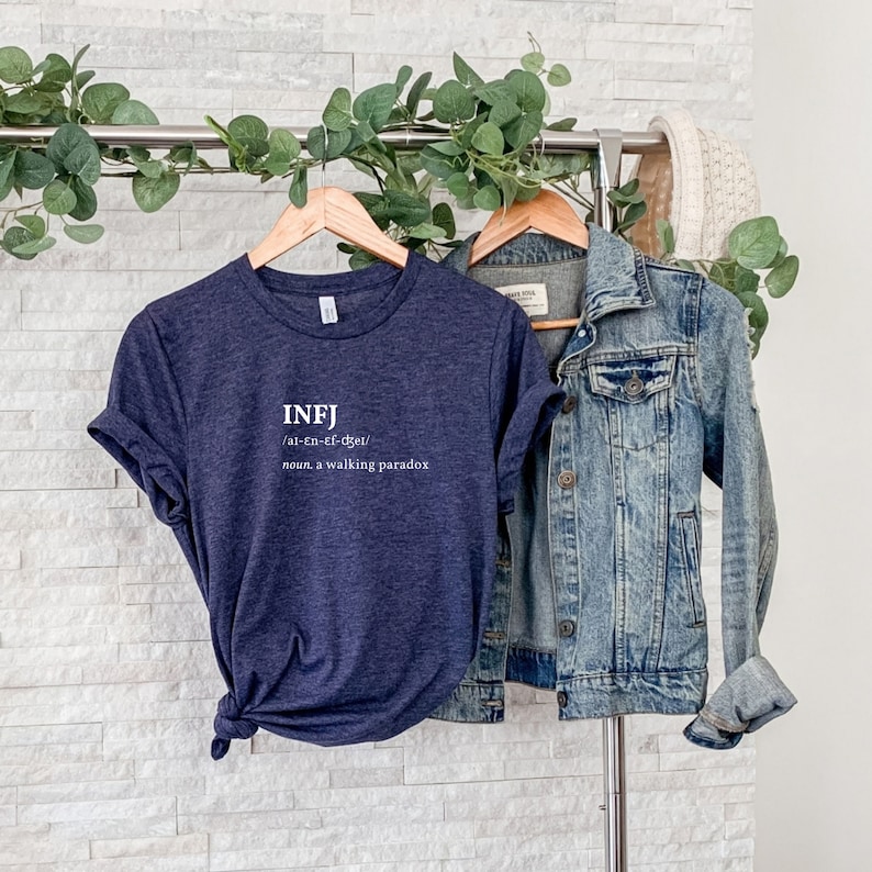 May include: A navy blue t-shirt with white text that reads "INFJ /ar-en-ef-djer/ noun. a walking paradox". The t-shirt is hanging on a wooden hanger with a denim jacket hanging next to it.