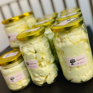 Pode incluir: Vários frascos de vidro cheios de um produto de manteiga batida de cor amarelo claro. Os frascos têm tampas douradas e etiquetas que dizem "Hairgrowth Butter" e "Dreamweaver Haircare".