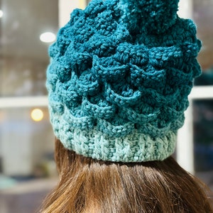 May include: A teal blue crocheted beanie with a fluffy pom pom on top. The beanie is made with a gradient yarn, starting with a darker teal blue and fading to a lighter teal blue.