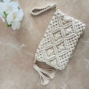 May include: A white crocheted clutch bag with a geometric pattern and a wooden bead tassel. The bag has a gold clasp and a woven strap.