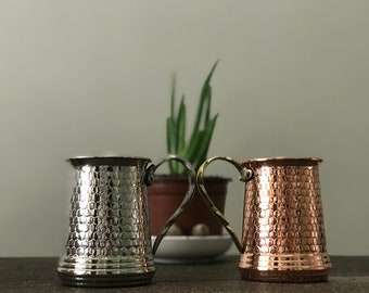 May include: Two decorative metal mugs with a honeycomb pattern. One mug is silver, the other is copper. Both have ornate handles. The mugs are likely for serving beverages.