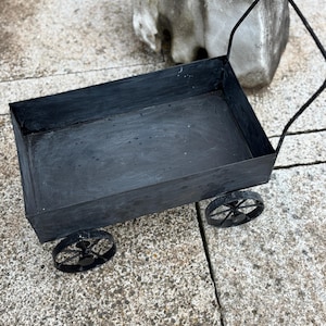 Könnte beinhalten: Ein kleiner, schwarzer Metallwagen mit rechteckiger Ladefläche und Griff. Der Wagen hat vier schwarze Speichenräder. Der Wagen steht auf einer Steinoberfläche.