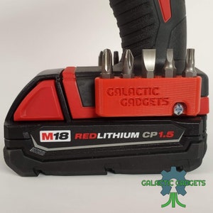 May include: A red and black power tool battery with the text "M18 REDLITHIUM CP1.5" and a red accessory holder with the text "GALACTIC GADGETS" attached to the top. The accessory holder has five slots for holding screwdriver bits.