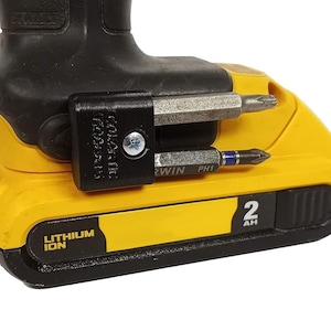 May include: A yellow and black power tool battery with a black plastic bit holder attached to the side. The bit holder has two screwdriver bits inserted. The battery has the text "LITHIUM ION" and "2 AH" printed on it.
