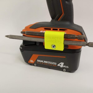 K&ouml;nnte beinhalten: Ein gelber 3D-gedruckter Werkzeughalter mit dem Text "GALACTIC GADGETS" befestigt an einem schwarzen und orangen Akku f&uuml;r ein Elektrowerkzeug. Zwei Metallbohrer werden im Halter gehalten.