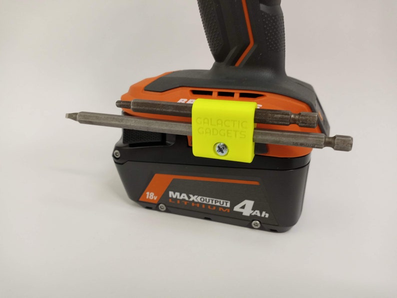 K&ouml;nnte beinhalten: Ein gelber 3D-gedruckter Werkzeughalter mit dem Text "GALACTIC GADGETS" befestigt an einem schwarzen und orangen Akku f&uuml;r ein Elektrowerkzeug. Zwei Metallbohrer werden im Halter gehalten.