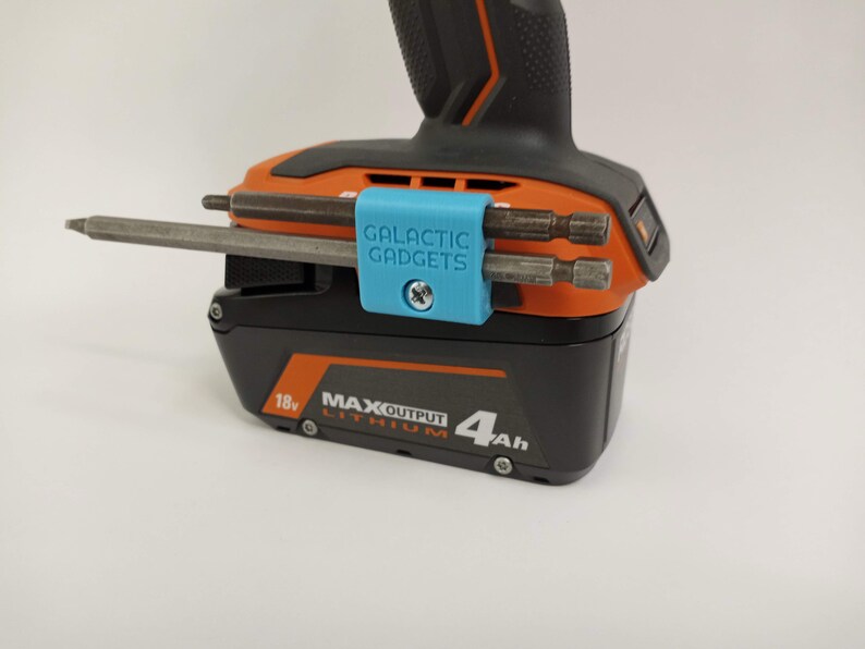 K&ouml;nnte beinhalten: Ein blauer 3D-gedruckter Werkzeughalter mit dem Text "GALACTIC GADGETS" befestigt an einem schwarzen und orangen Akkuschrauber-Akku. Der Halter h&auml;lt drei unterschiedlich gro&szlig;e Bohrer.