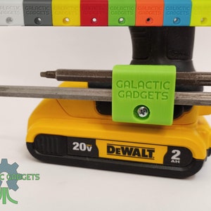 May include: A green 3D printed tool holder with the text "GALACTIC GADGETS" attached to a yellow DeWalt 20V MAX battery. The holder is designed to hold two screwdriver bits.