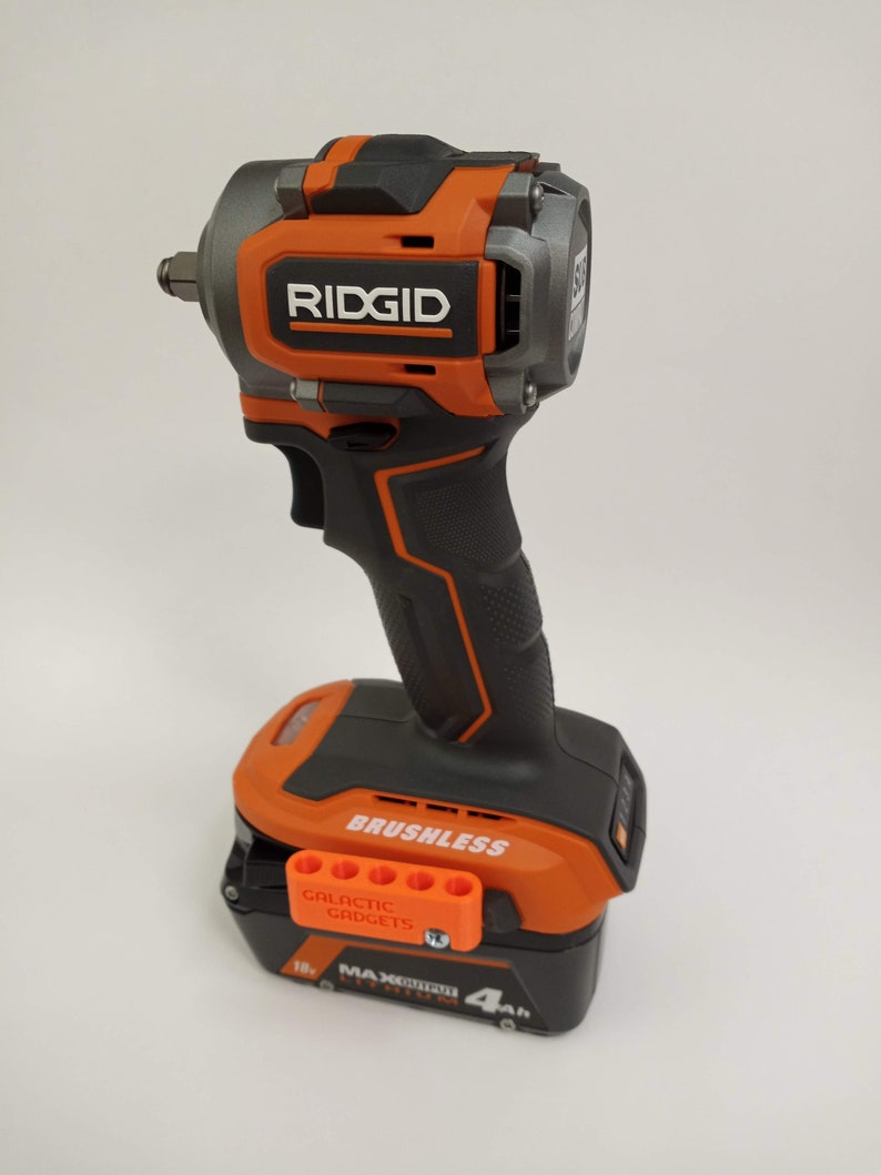 May include: An orange and black cordless impact wrench with a brushless motor and a 4Ah battery. The battery has a label that says "18V MAX OUTPUT LITHIUM ION 4Ah". The wrench has a black handle and a silver head. The wrench is made by RIDGID.