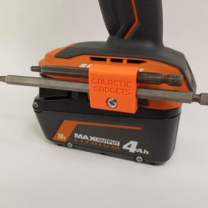 K&ouml;nnte beinhalten: Orangefarbener 3D-gedruckter Werkzeughalter mit dem Text "GALACTIC GADGETS", der an einem schwarzen und orangen Akku f&uuml;r Elektrowerkzeuge befestigt ist. Der Halter hat zwei Metallbohrer, die daran befestigt sind.