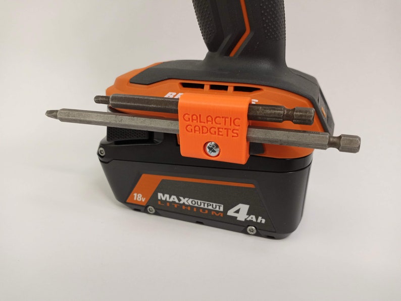 K&ouml;nnte beinhalten: Orangefarbener 3D-gedruckter Werkzeughalter mit dem Text "GALACTIC GADGETS", der an einem schwarzen und orangen Akku f&uuml;r Elektrowerkzeuge befestigt ist. Der Halter hat zwei Metallbohrer, die daran befestigt sind.