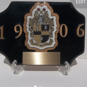 Puede incluir: Una placa negra con letras doradas y un escudo dorado y negro. La placa dice "1906" y presenta un escudo con las palabras "Alpha Phi Alpha".