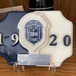 May include: A white and blue ceramic plaque with a crest featuring the letters "ZΦΒ" and the year "1920". The plaque has the years "19" and "20" on either side of the crest. The plaque has a silver metal plate with the words "Pure Aspiration" on it.