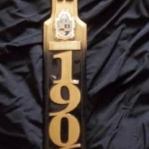 May include: A black and gold wooden paddle with the letters "AKA" and the year "1906" engraved on it. The paddle has a small crest with the letters "AKA" and the number "19" on it.