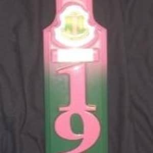 Peut inclure: Une pagaie en bois rose et verte avec les lettres "AKA" et l'année "1908" imprimées dessus. La pagaie a une petite plaque en bas avec les mots "Alpha Kappa Alpha Sorority, Incorporated" imprimés dessus.