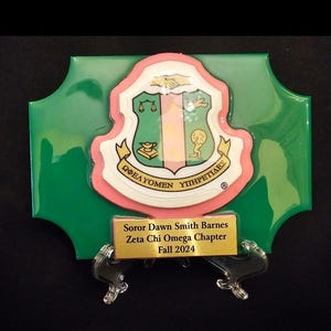 May include: A green and pink sorority plaque with a gold inscription that reads "Soror Dawn Smith Barnes Zeta Chi Omega Chapter Fall 2024".