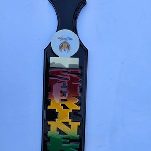 May include: A black wooden paddle with a gold and red "Shriner" inscription. The paddle has a white circle with a gold and red Shriner emblem on it.